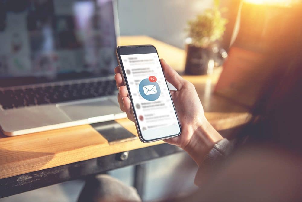 Vor einem Monitor ist ein Smartphone zu sehen, auf dessen Bildschrim ein E-Mail Newsletter eingegangen ist