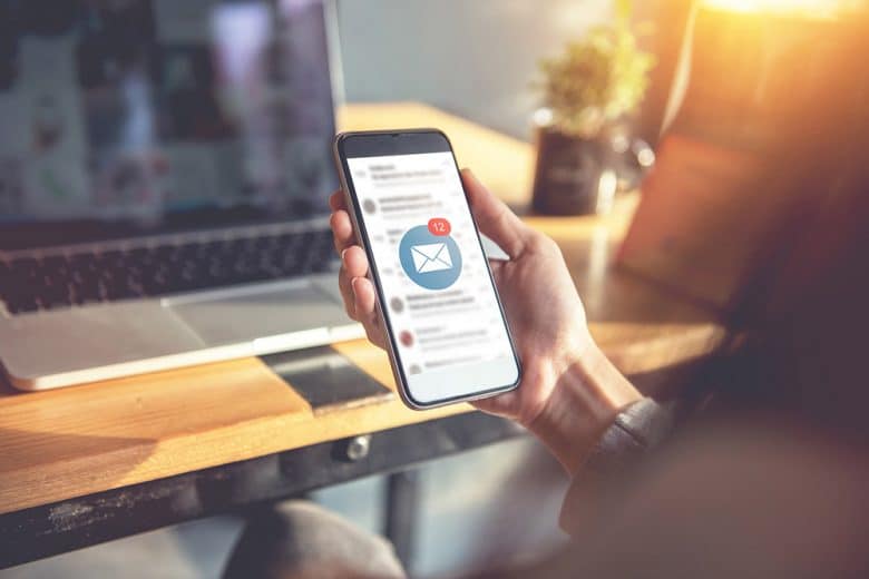 Vor einem Monitor ist ein Smartphone zu sehen, auf dessen Bildschrim ein E-Mail Newsletter eingegangen ist