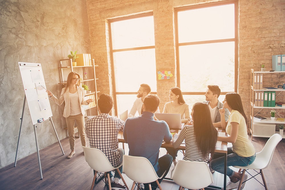 Eine junge Frau präsentiert an einem Flipchart vor 7 weiteren jungen Menschen wovon einer einen Laptop nutzt