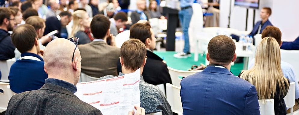 Man sieht viele Leute auf Stühlen sitzen bei einer Veranstaltung, die einem Vortragenden zuhören