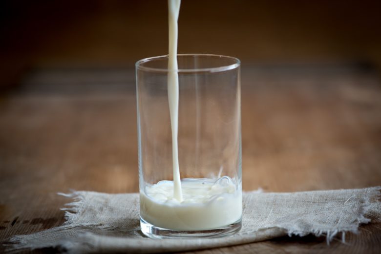 In ein Glas wird Milch eingeschenkt. Es steht auf einem braunen Holztisch